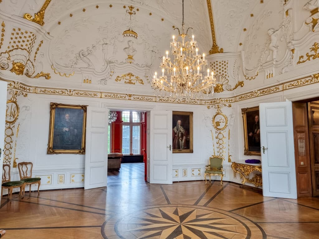 Weißer Saal mit kunstvoll verzierten Wänden, Decken mit weiß und Gold sowie Gemälden an den Wänden und Holzboden