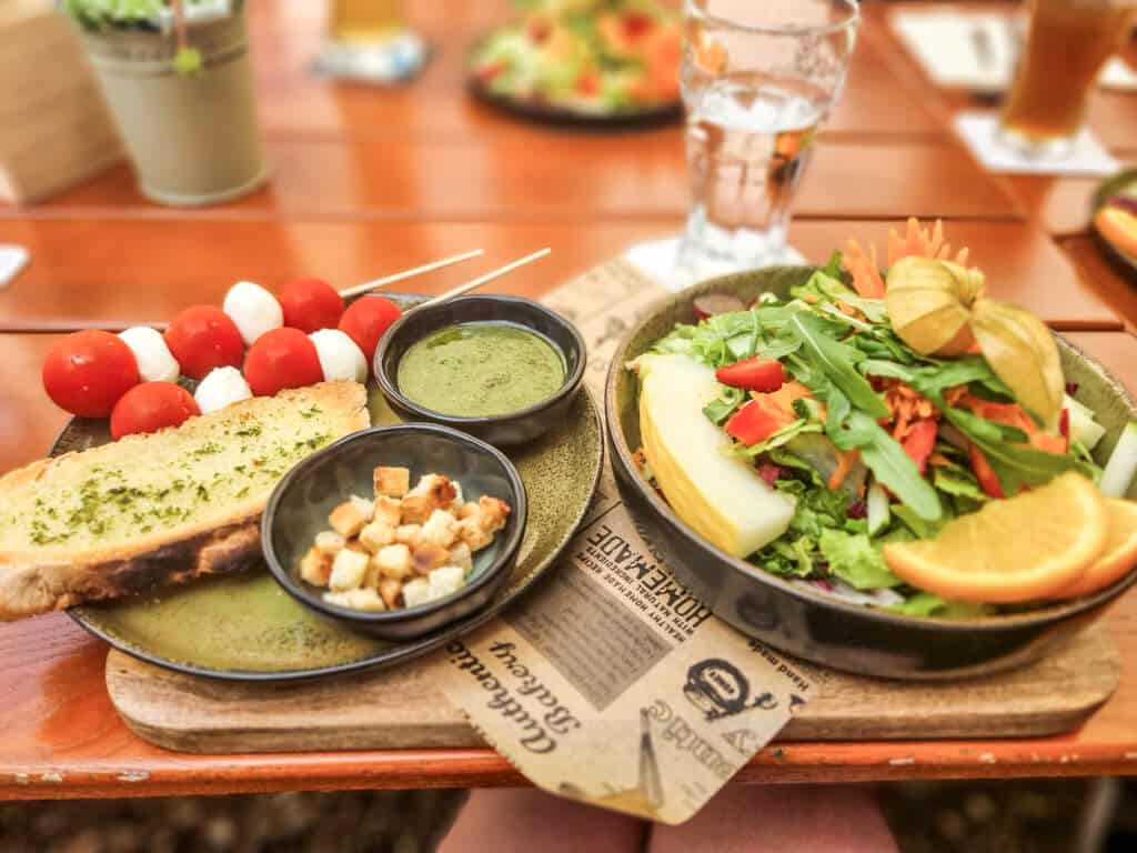 Ein Teller mit frischem Salat, garniert mit Orangenscheiben, Kirschtomaten, Blattgemüse und anderem bunten Gemüse. Daneben ein Stück Kräuterbrot, ein Schälchen mit Croutons, eine grüne Dip-Sauce und Spieße mit Mozzarellakugeln und Kirschtomaten.