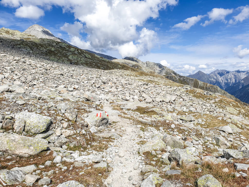 schmaler Weg mit weiß-roter Markierung für den Wanderweg führt zwischen Geröllfeldern über einen Bergkamm. Im Hintergrund sind hohe Berge bei blauem Himmel zu sehen