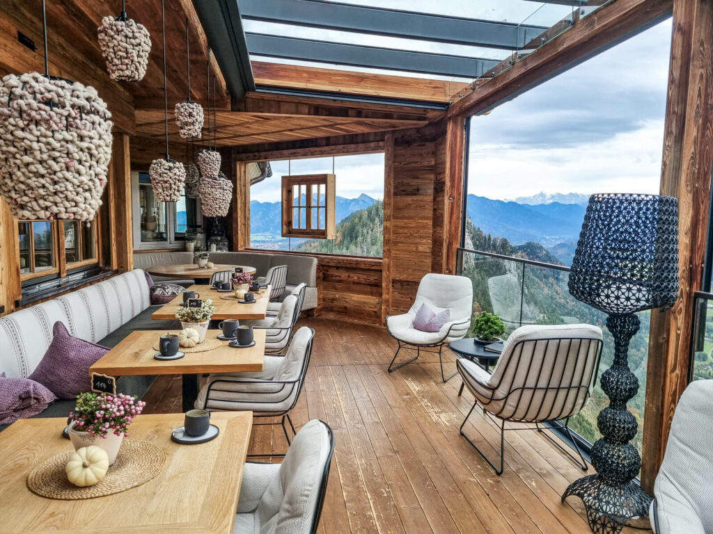 Ein gemütliches Café mit Bergblick, Holzinterieur, Plüschsitzen und dekorativen Hängelampen. Die Tische sind mit Tassen und kleinen Blumen gedeckt. Große Fenster bieten einen malerischen Blick auf die fernen Berge unter einem bewölkten Himmel.
