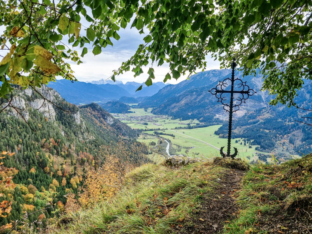 Eine malerische Aussicht von einem Hügel, eingerahmt von Ästen, auf ein üppiges grünes Tal. Im Vordergrund steht ein dekoratives Metallkreuz, in der Ferne sind Berge unter einem teilweise bewölkten Himmel zu sehen.
