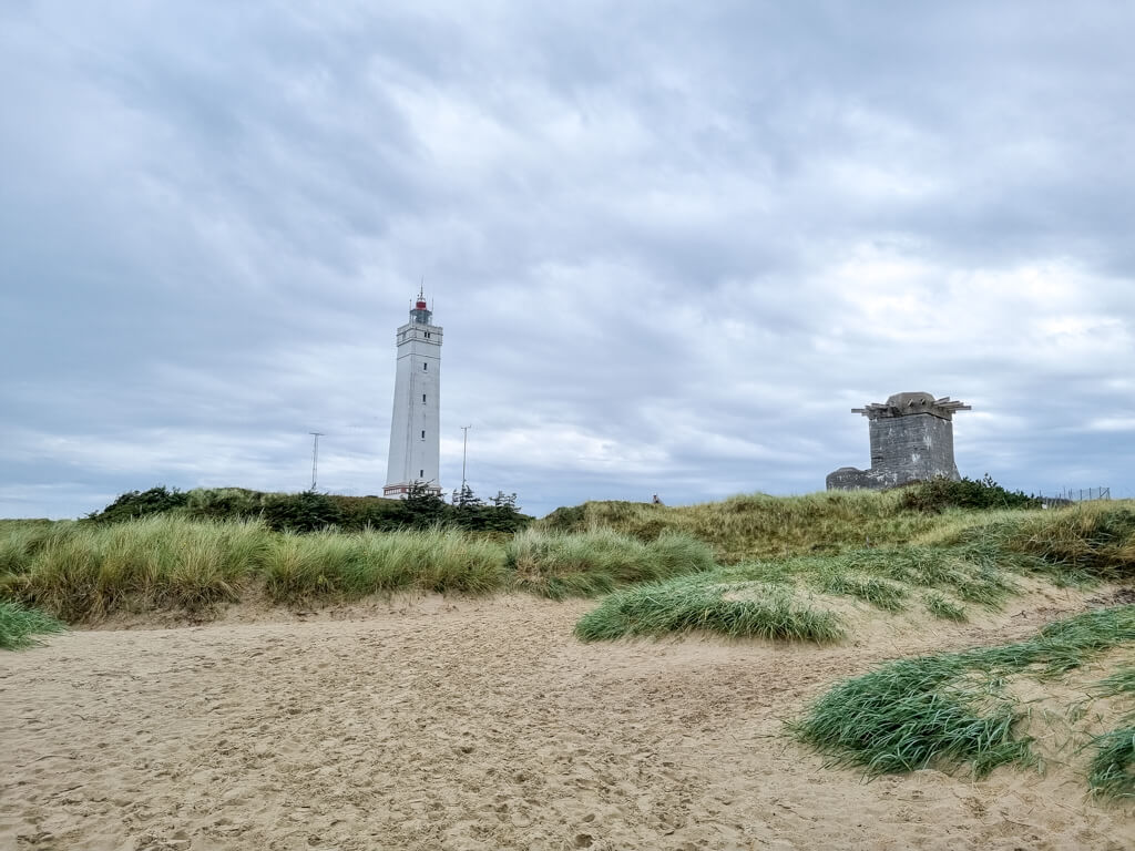 weißer Leuchtturm inmitten von grasbewachsenen Dünen; davor ein heller Sandstrand; rechts neben dem Leuchtturm befindet sich ein Bunker