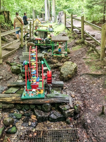 Zwei Menschen gehen einen Waldweg entlang, der an farbenfrohen Modellen von Wasserrädern vorbeiführt. Im Vordergrund ist ein durch Wasserkraft betriebenes Riesenrad zu sehen. Die Gegend ist üppig grün und ein Holzzaun säumt den Weg.
