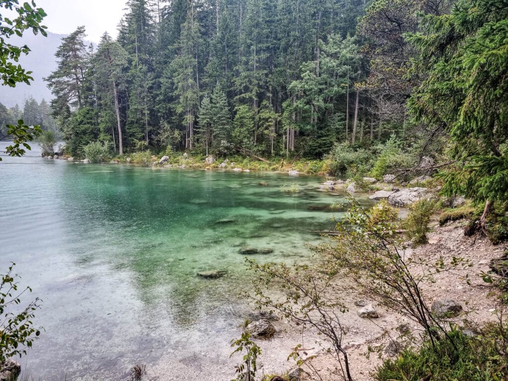 Eine ruhige Szene am Seeufer mit klarem türkisfarbenem Wasser und einem sandigen Ufer. Dichte immergrüne Bäume säumen den See und bilden eine ruhige Waldkulisse unter einem bewölkten Himmel.
