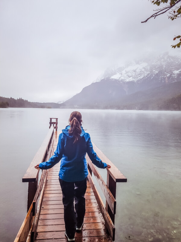 Eine Person in einer blauen Jacke steht auf einem Holzsteg und blickt auf einen nebligen See mit schneebedeckten Bergen im Hintergrund. Der Himmel ist bedeckt, was eine ruhige Atmosphäre schafft.
