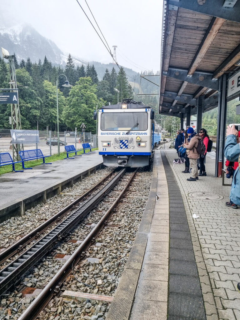 Ein blau-weißer Zug mit der Aufschrift „Bayerische Zugspitzbahn“ steht auf einem Bahnsteig, umgeben von Menschen. Der Bahnhof bietet eine Bergkulisse mit Bäumen und bewölktem Himmel. Die Passagiere sind für kühles Wetter gekleidet.
