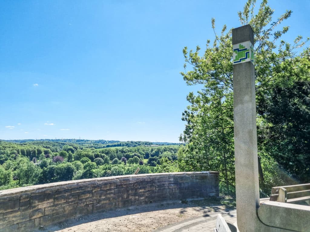 Ein Steinpfosten mit einem grün-blauen Schild steht neben einer geschwungenen Steinmauer mit Blick auf einen üppigen, grünen Wald unter einem klaren blauen Himmel.