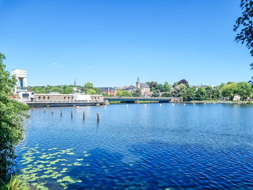 Ein ruhiger See mit Seerosen im Vordergrund, Gebäuden und einer Kirche mit einem hohen Kirchturm am Ufer und Bäumen unter einem klaren blauen Himmel.

