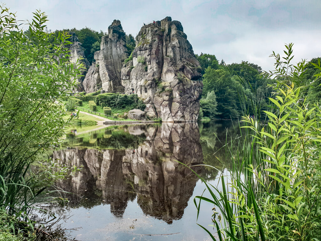 Externsteine spiegeln sich im darunter liegenden See. Im Vordergrund sind einige Gräser sichtbar