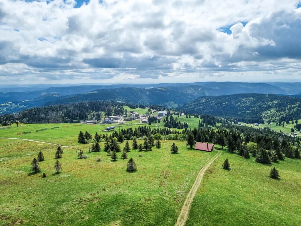 Eine malerische Luftaufnahme von sanften grünen Hügeln mit Kiefern, einem Feldweg, der zu einem Gebäude mit rotem Dach führt, und einer Ansammlung von Gebäuden im Hintergrund unter einem teilweise bewölkten Himmel.