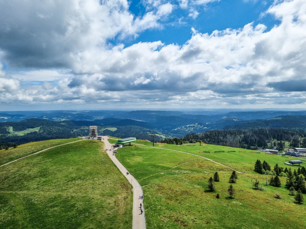 Eine malerische Aussicht auf sanfte grüne Hügel unter einem teilweise bewölkten Himmel, mit einem gewundenen Pfad, vereinzelten Bäumen, einigen Gebäuden und in der Ferne Wälder und Berge am Horizont.