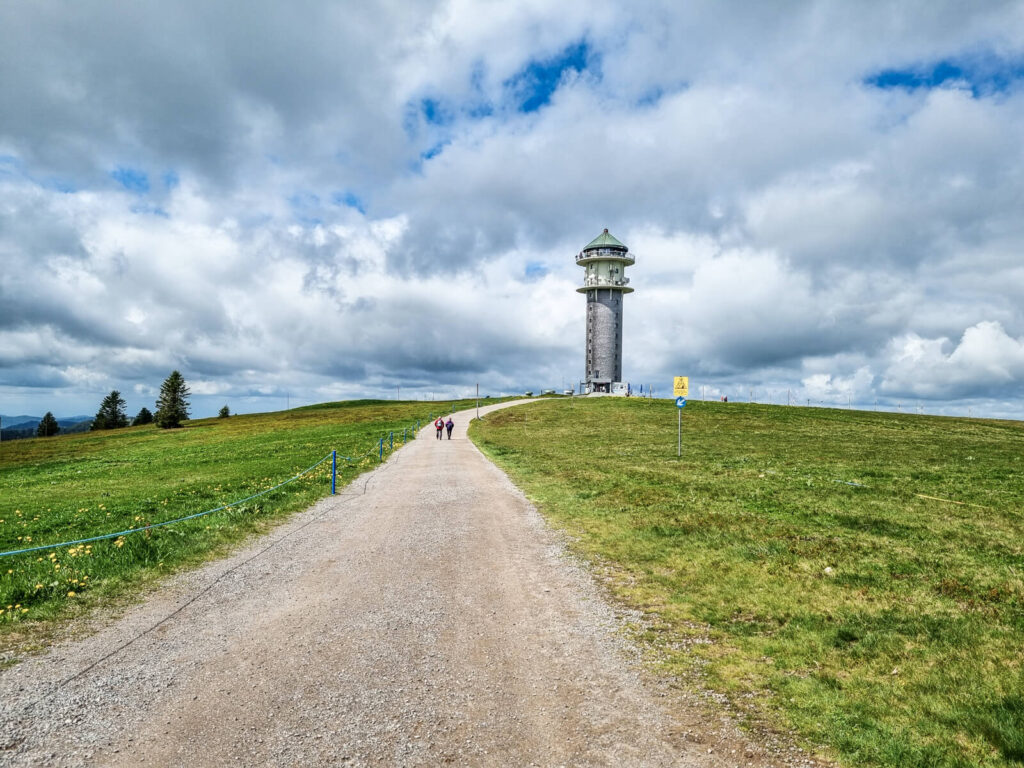 Ein Schotterweg führt unter einem teilweise bewölkten Himmel einen grasbewachsenen Hügel hinauf zu einem hohen Aussichtsturm. Ein paar Menschen gehen den Weg entlang.

