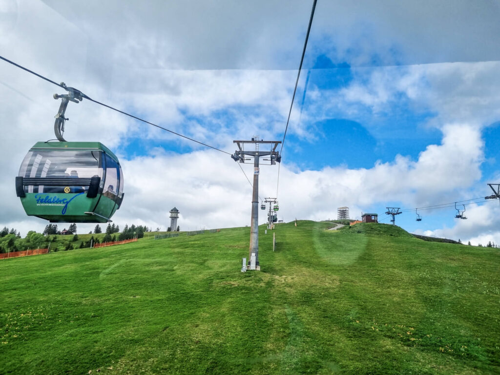 Eine grüne Seilbahn fährt unter blauem, bewölktem Himmel einen grasbewachsenen Hügel hinauf. Metalltürme stützen die Seilbahn, in der Ferne sind weitere Seilbahnen und ein Gebäude zu sehen.