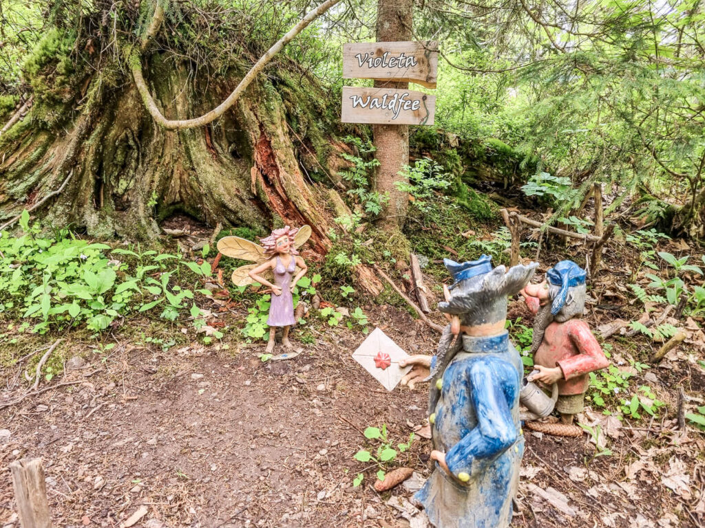 Drei Statuen (Fee und zwei Wichtel) stehen auf einer Waldlichtung in der Nähe eines Baumes mit Holzschildern mit der Aufschrift „Violeta Waldfee“. Wichtelpostbote hält einen Umschlag in der Hand. Die Umgebung ist üppig grün und mit Baumwurzeln übersät.

