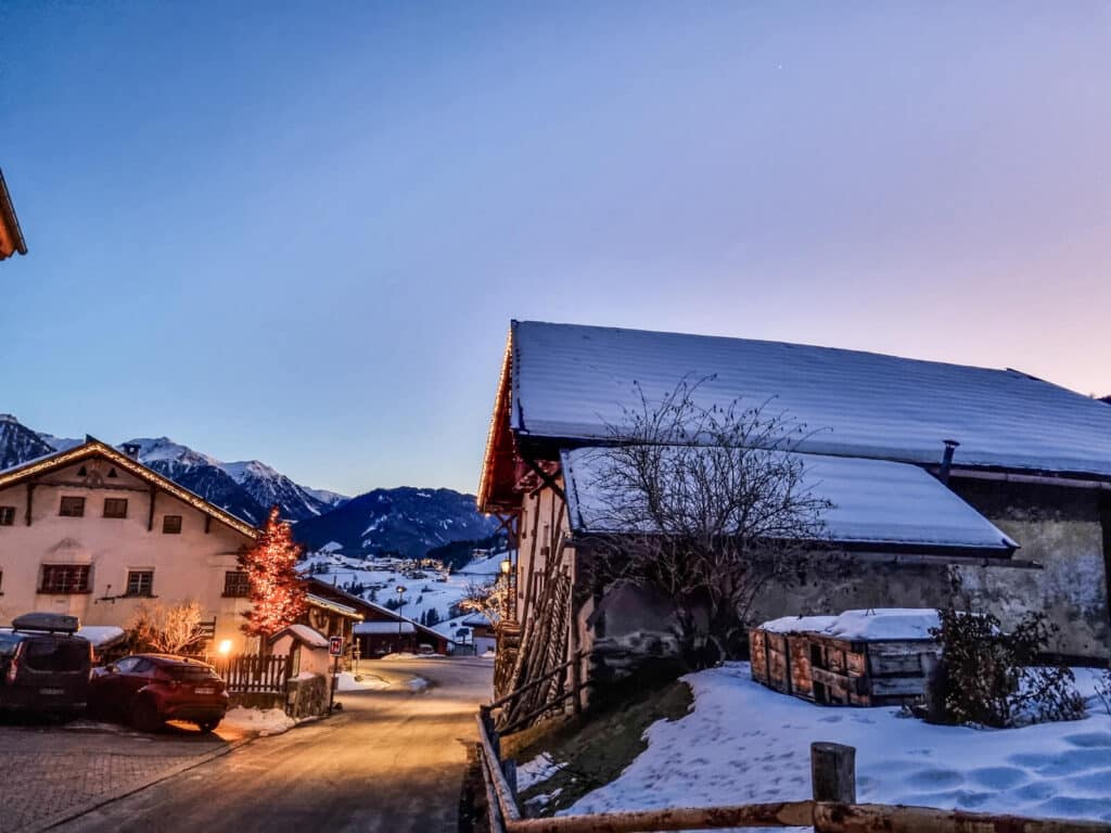 Schneebedecktes Dorf in der Abenddämmerung mit einem Baum, der von roten Lichtern bedeckt ist. Häuser und Dächer sind mit Schnee bedeckt, und im Hintergrund sind Berge unter einem klaren Abendhimmel zu sehen. Ein Auto ist in der Nähe des Baumes geparkt.
