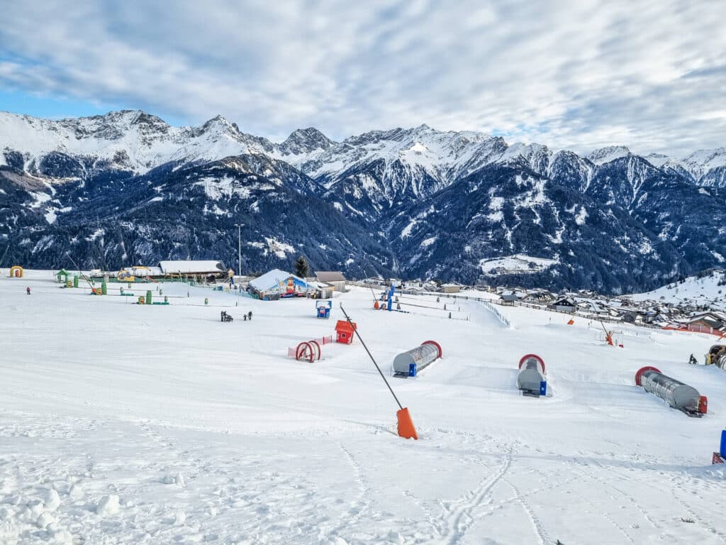 Verschneite Berglandschaft mit Skipisten und Förderbändern für Kinder im Vordergrund, auf denen verschiedene Skiausrüstungen und -strukturen zu sehen sind. Im Hintergrund sind schneebedeckte Gipfel unter einem teilweise bewölkten Himmel zu sehen.
