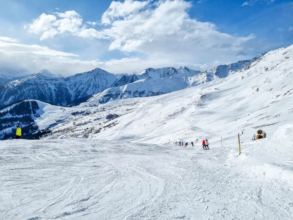 Verschneite Berglandschaft mit klarem, blauem Himmel. Skifahrer verteilen sich über den Hang, im Hintergrund sind Berggipfel zu sehen. Ein paar Wolken zieren den Himmel und verleihen der ausgedehnten Winterszene Tiefe.

