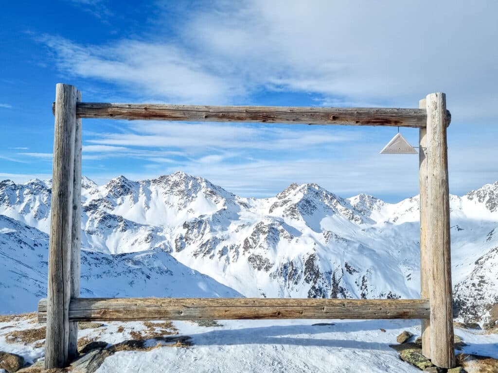 Holzrahmen auf einem schneebedeckten Berggipfel, der einen Panoramablick auf entfernte schneebedeckte Gipfel unter einem teilweise bewölkten blauen Himmel bietet. An der oberen rechten Ecke des Rahmens hängt eine kleine dreieckige Holzdekoration.
