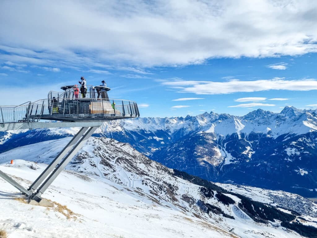 Eine moderne Aussichtsplattform mit mehreren Personen bietet einen Blick auf schneebedeckte Berge und Täler unter einem teilweise bewölkten Himmel. Die Landschaft zeichnet sich durch schroffe Gipfel und ausgedehnte schneebedeckte Flächen aus und schafft eine malerische Winterszene.
