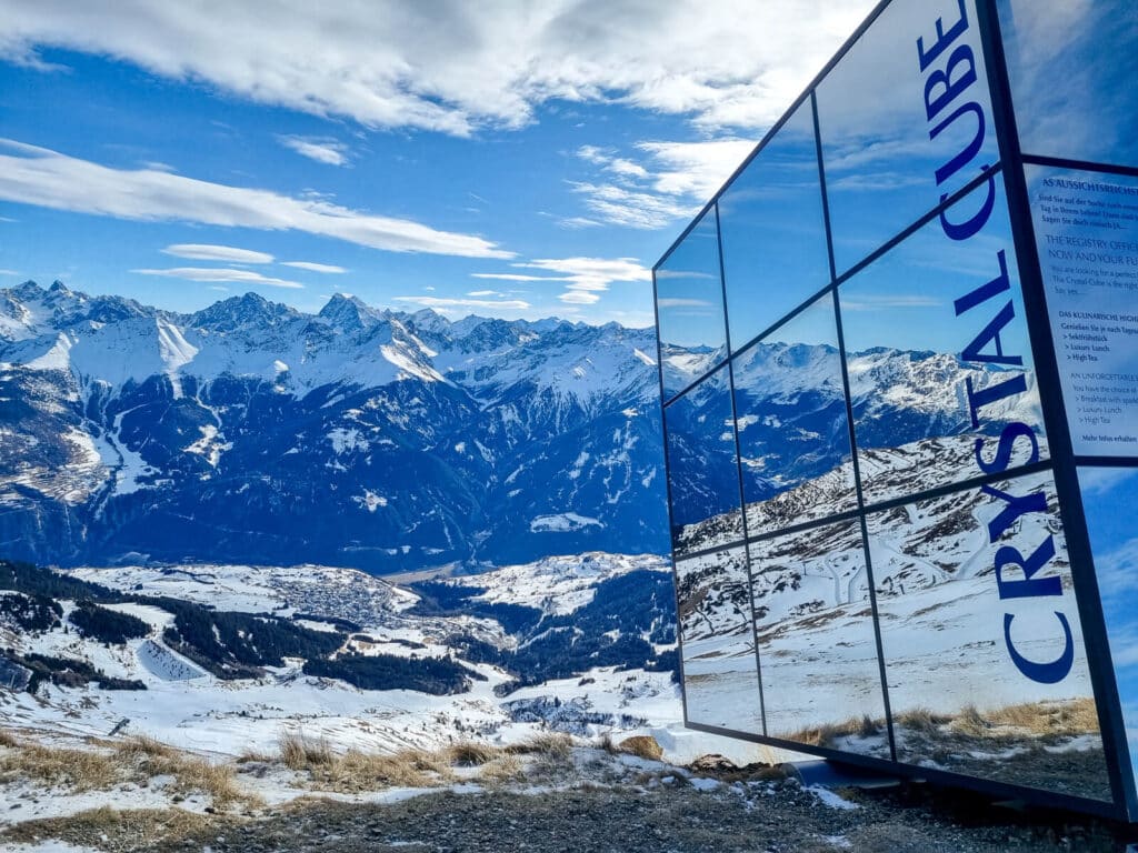 Ein verspiegeltes Gebäude mit der Aufschrift „Crystal Cube“ reflektiert eine atemberaubende schneebedeckte Bergkette unter einem teilweise bewölkten blauen Himmel. Die Landschaft zeichnet sich durch schneebedeckte Täler und Gipfel aus, die ein ruhiges Alpenpanorama erzeugen.
