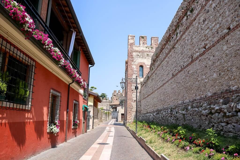 Stadtmauer auf der rechten Seite, links ein rot gestrichenes Haus mit Blumen