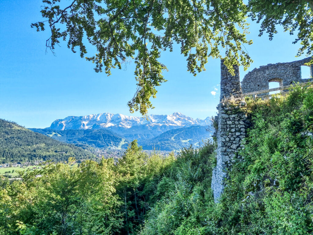 Alte Steinruinen sind von üppigem Grün umgeben, vor der Kulisse majestätischer, schneebedeckter Berge unter einem klaren blauen Himmel. Sonnenlicht fällt durch die Äste der Bäume und schafft eine ruhige und malerische Landschaft.