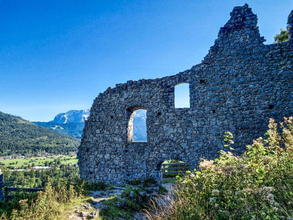 Steinmauer eines historischen Gebäudes (Ruine) mit drei Fensteröffnungen vor der Kulisse grüner Hügel und ferner Berge unter einem klaren blauen Himmel. Im Vordergrund neben der Mauer wachsen wilde Pflanzen.