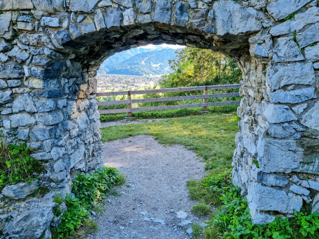 Ein Steinbogen führt zu einem Grasweg und einem Holzzaun. Im Hintergrund blickt man auf die Berge. Üppiges Grün umgibt den Weg, und der Himmel ist hell und klar.