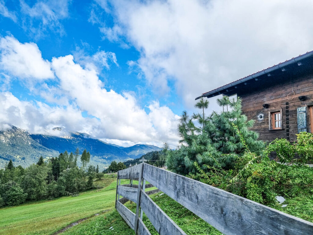 Rustikale Holzhütte in der Nähe einer grünen Wiese und Kiefern, mit einem Holzzaun im Vordergrund. Unter einem teilweise bewölkten blauen Himmel sind Berge sichtbar.