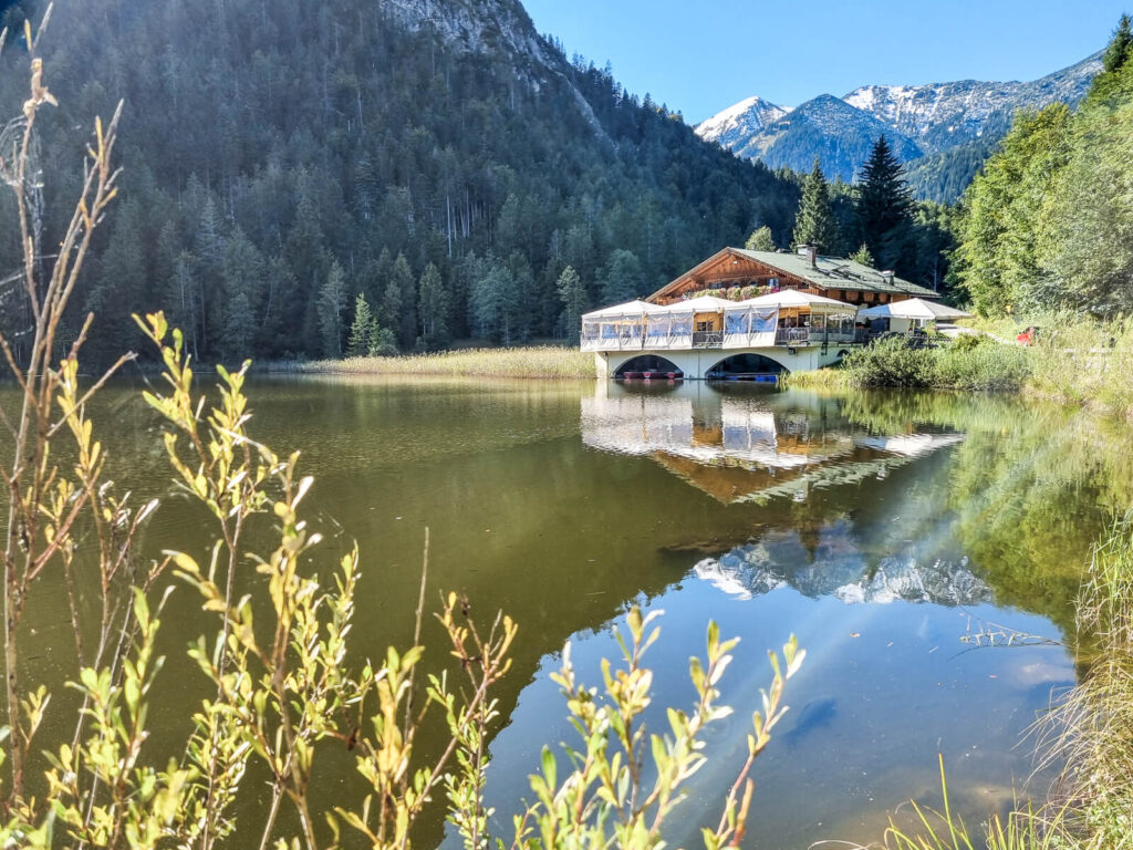 Ein ruhiger See mit einem Pfahlbau in Ufernähe, umgeben von üppigen grünen Bäumen und Bergen im Hintergrund. Das Wasser spiegelt das Bauwerk und die Berge. Helles Sonnenlicht hebt das Laub im Vordergrund hervor.