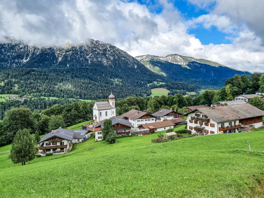 Ein malerisches Dorf in einem üppig grünen Tal mit traditionellen Alpenhäusern, einer markanten weißen Kirche mit Turm und umgeben von bewaldeten Hügeln und schneebedeckten Bergen unter einem teilweise bewölkten Himmel.