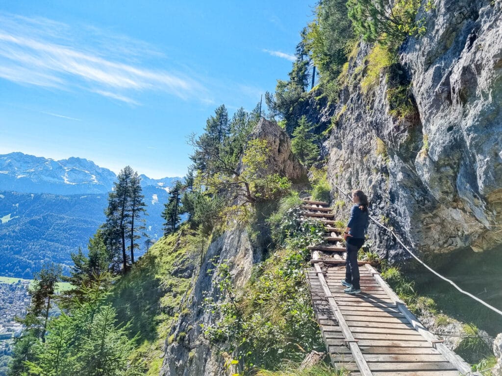 Eine Person steht auf einem schmalen Holzsteg, der in einen felsigen Berghang gebaut ist, und genießt den Panoramablick auf die fernen Berge und das darunterliegende Tal. Der Himmel ist klar und blau, und die Gegend ist üppig grün.

