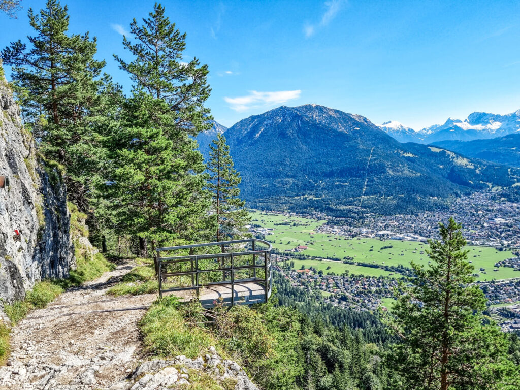 Ein malerischer Blick auf eine Berglandschaft mit einem felsigen Pfad, der zu einem Aussichtspunkt mit Metallgeländer führt. Immergrüne Bäume säumen den Weg. Darunter ist ein grünes Tal mit einer Stadt zu sehen, dahinter eine große Bergkette unter einem klaren blauen Himmel.