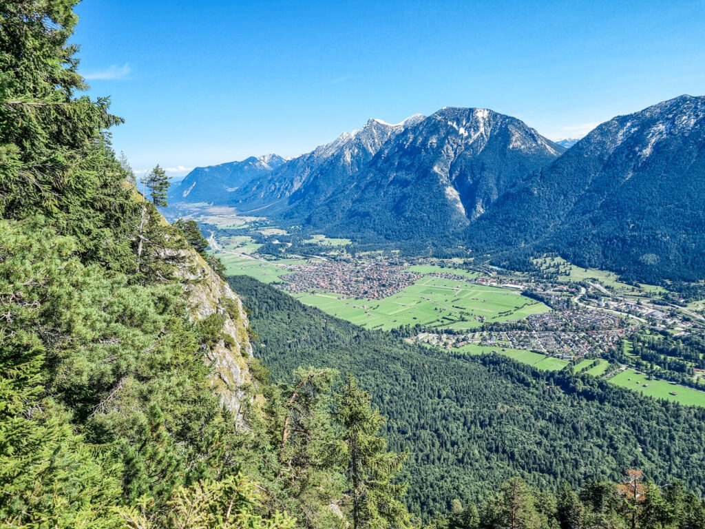 Ein malerischer Blick auf Berge mit schneebedeckten Gipfeln und ein üppiges Tal darunter. Das Tal ist mit einer kleinen Stadt und Feldern übersät, umgeben von dichten Wäldern unter einem klaren blauen Himmel.