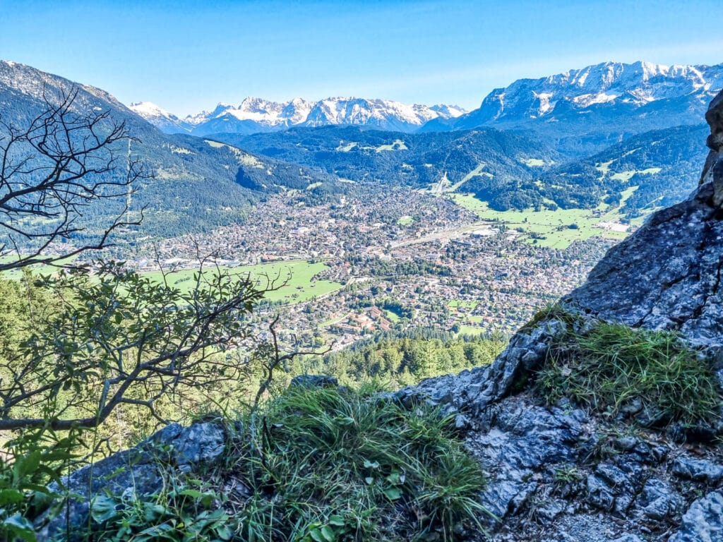 Malerischer Blick von einem Felsvorsprung auf eine üppig grüne Talstadt, umgeben von Bergen unter klarem blauen Himmel. Auf den Felsen im Vordergrund wächst spärliche Vegetation. In der Ferne sind schneebedeckte Gipfel sichtbar.
