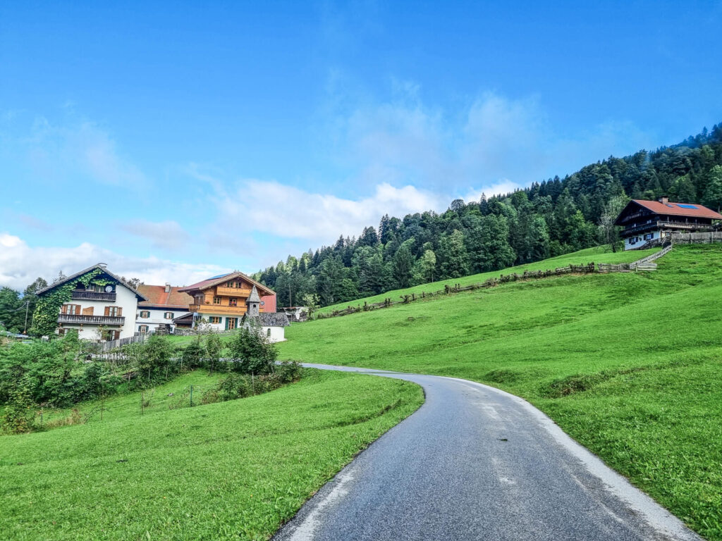 Eine kurvenreiche Straße führt durch üppige, grüne Hügel zu einer Ansammlung von Häusern mit roten Dächern. Dichte Bäume säumen die Szenerie, und der strahlend blaue Himmel mit einigen Wolken schafft eine malerische Landschaftskulisse.