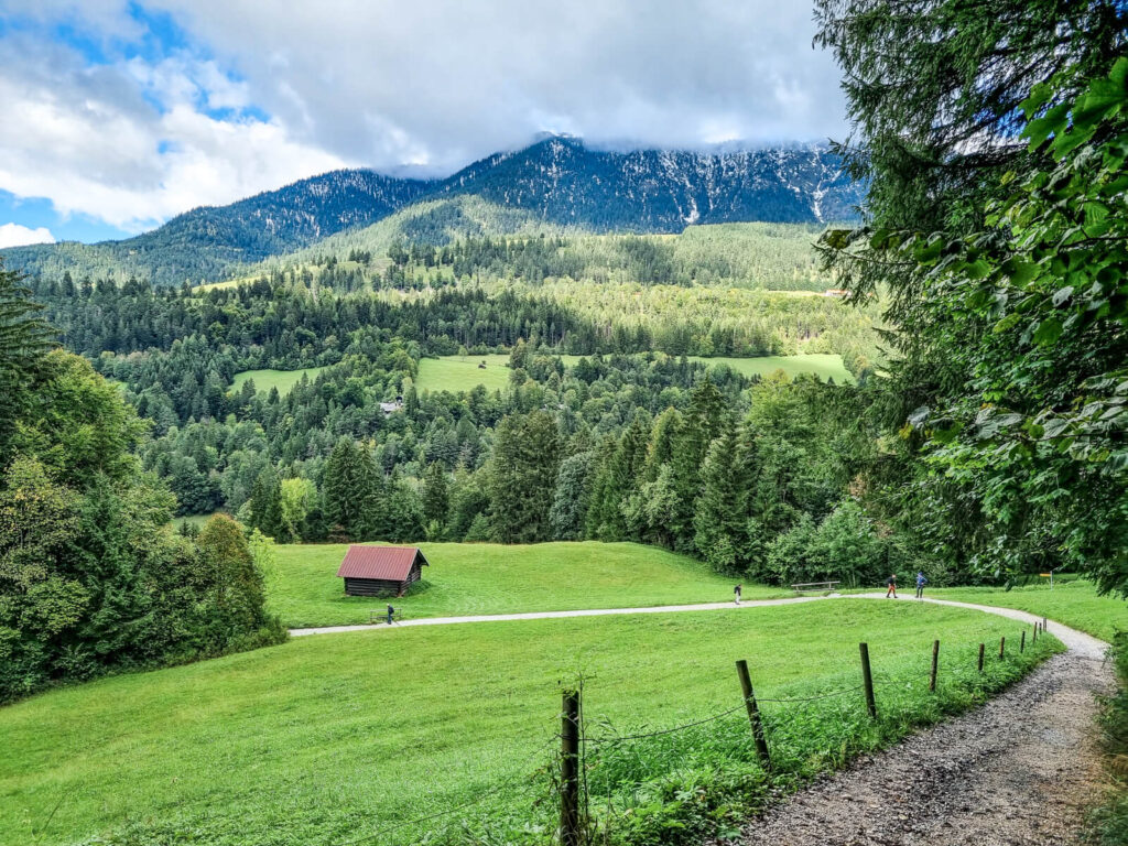 Malerische Landschaft mit einem Feldweg, der sich durch eine grüne, von einem Zaun gesäumte Wiese schlängelt. Eine kleine Hütte mit rotem Dach liegt eingebettet zwischen den Bäumen. Dichte Wälder bedecken die sanften Hügel, im Hintergrund sind unter einem teilweise bewölkten Himmel Berggipfel zu sehen.