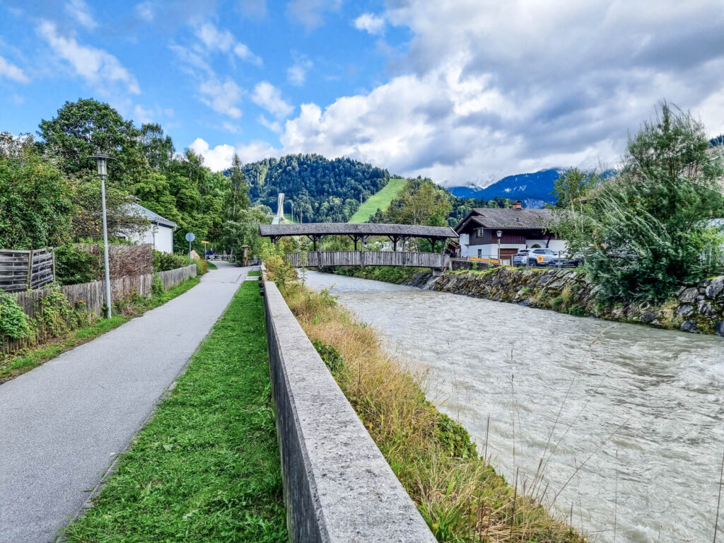 Weg entlang eines Flusses mit üppigem Grün und einer Holzbrücke. Im Hintergrund sind Häuser und Berge unter einem teilweise bewölkten Himmel zu sehen.