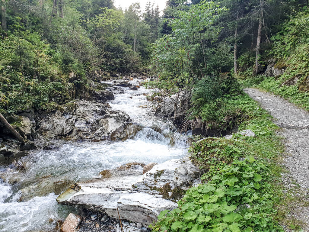 Fluss fließt stufenförmig über einige Felsen und Steine; rechts davon ein kleiner Weg und Gräser