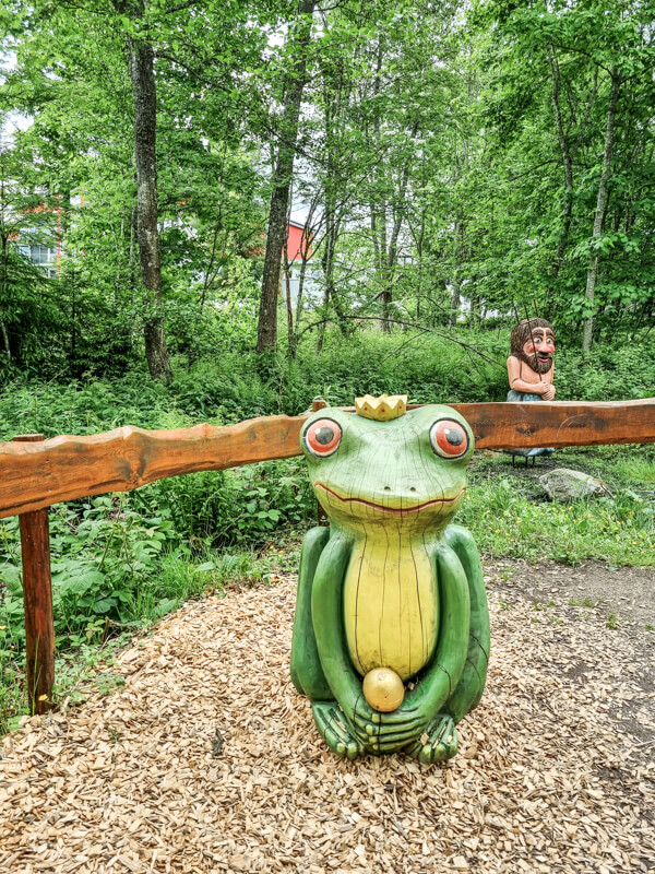 Eine große, farbenfrohe Froschstatue aus Holz mit einer Krone steht im Freien in einem Waldgebiet vor einem rustikalen Holzzaun. Im Hintergrund ist zwischen grünen Bäumen eine bärtige Figur zu sehen.