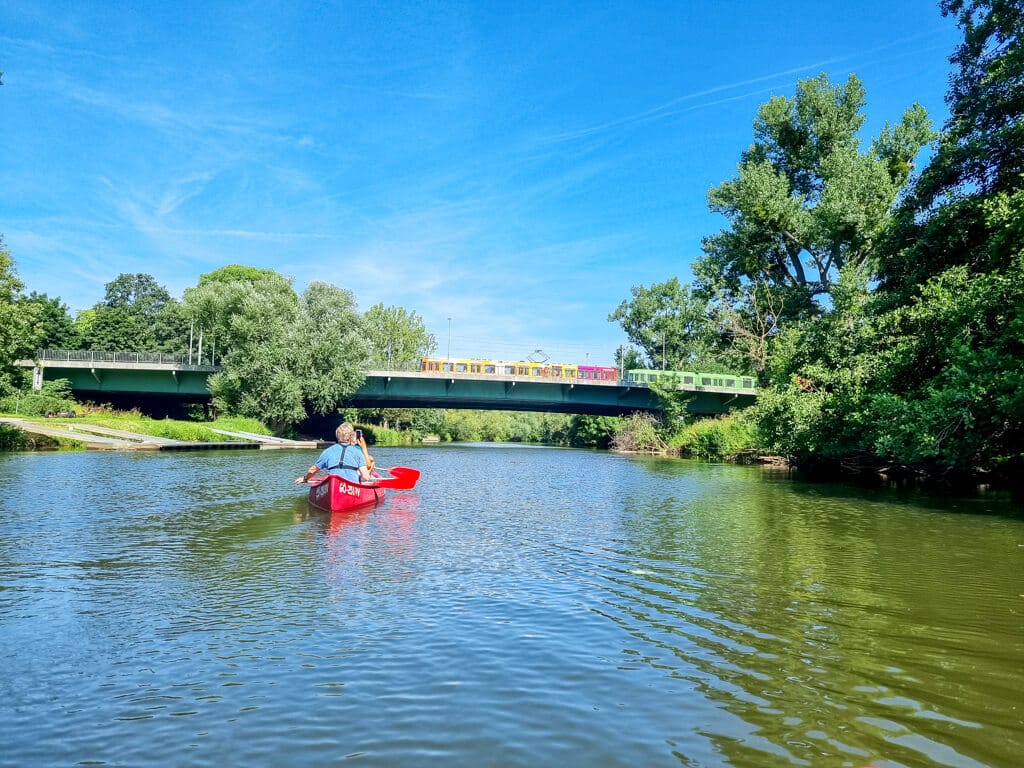 Eine Person paddelt in einem roten Kanu auf einem ruhigen Fluss, umgeben von üppigen grünen Bäumen unter einem klaren blauen Himmel. Im Hintergrund überspannt eine Brücke, über die eine S-Bahn mit farbenfrohen Wagen fährt, den Fluss.
