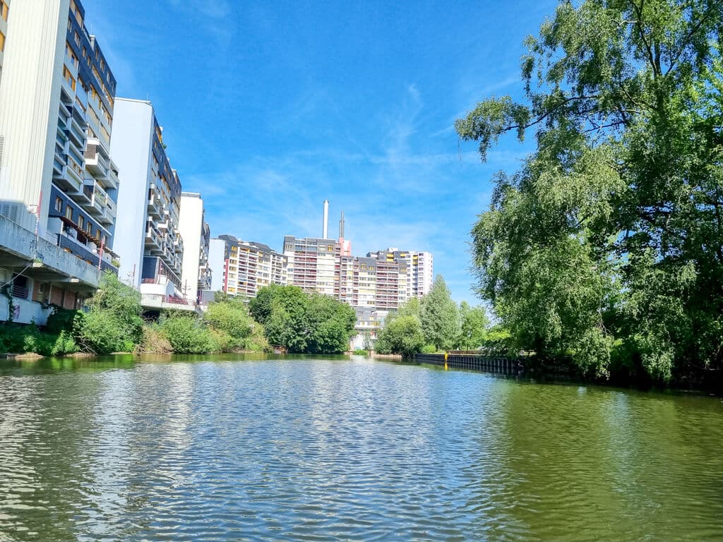 Ein malerischer Blick auf einen ruhigen Fluss, gesäumt von üppigen grünen Bäumen und hohen Plattenbauten unter einem klaren blauen Himmel. Das Wasser spiegelt das Grün und die Gebäude wider und schafft eine ruhige Stadtlandschaft.
