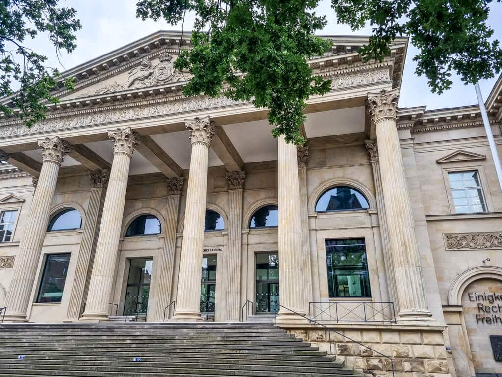 Das Bild zeigt die Fassade eines großen neoklassizistischen Gebäudes mit sechs hohen Säulen und einem verzierten Giebel. Das Gebäude hat große, gewölbte Fenster und eine Reihe breiter Stufen, die zum Eingang hinaufführen. Im Vordergrund sind grüne, belaubte Zweige zu sehen.