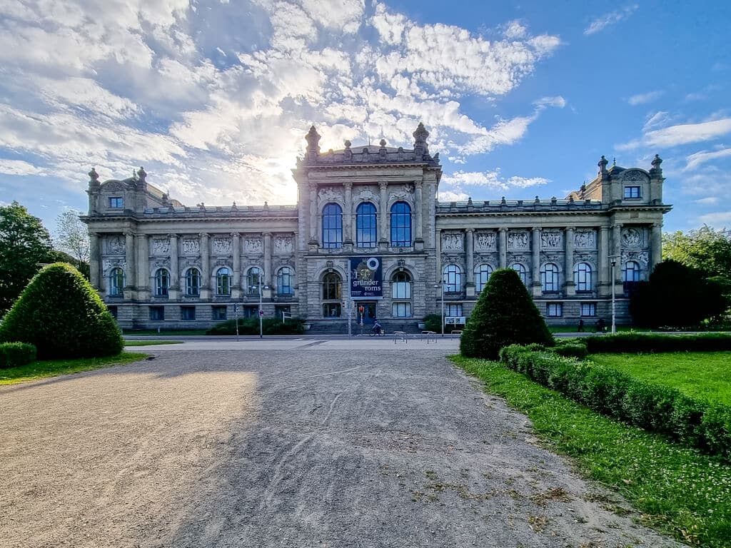 Ein großes, verziertes historisches Gebäude mit detailliertem Mauerwerk und Rundbogenfenstern, umrahmt von gestutzten Hecken und einem Kiesweg unter einem teilweise bewölkten Himmel.
