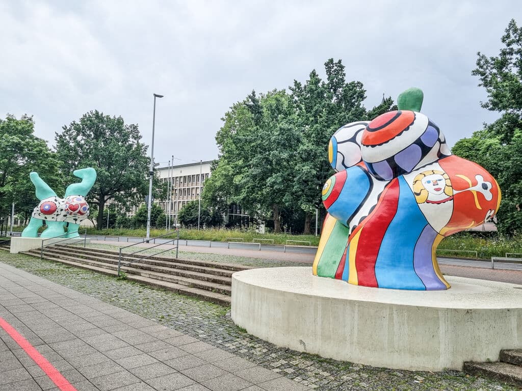 Bunte, abstrakte Skulpturen werden im Freien auf großen Betonplatten ausgestellt. Die Skulptur im Vordergrund weist helle Muster und menschenähnliche Gesichter auf, während die Skulptur im Hintergrund grün mit roten und weißen Akzenten ist. Beide stehen vor Bäumen und einem Gebäude.
