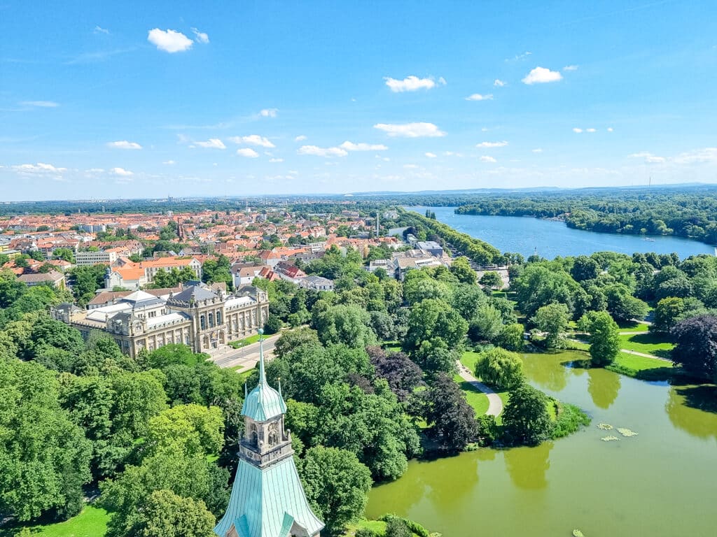 Ausblick über zwei Seen mit vielen Bäumen rundherum. Linksseitig ein historisches Gebäude mit Säulen; dahinter eine Stadt