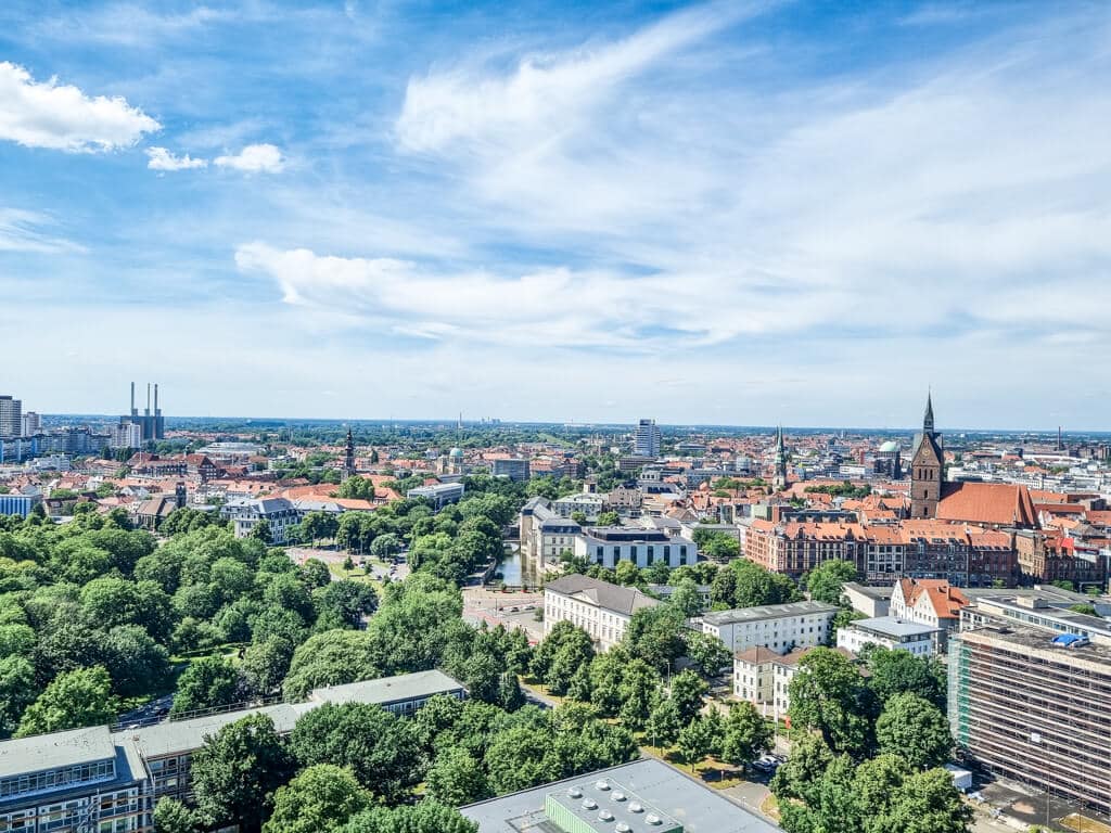 Luftaufnahme einer Stadt mit einer Mischung aus moderner und historischer Architektur. Zu den markanten Gebäuden gehören eine hohe Kirche mit rotem Dach und ein großer Park mit vielen grünen Bäumen im Vordergrund. Der Himmel darüber ist klar mit vereinzelten Wolken.
