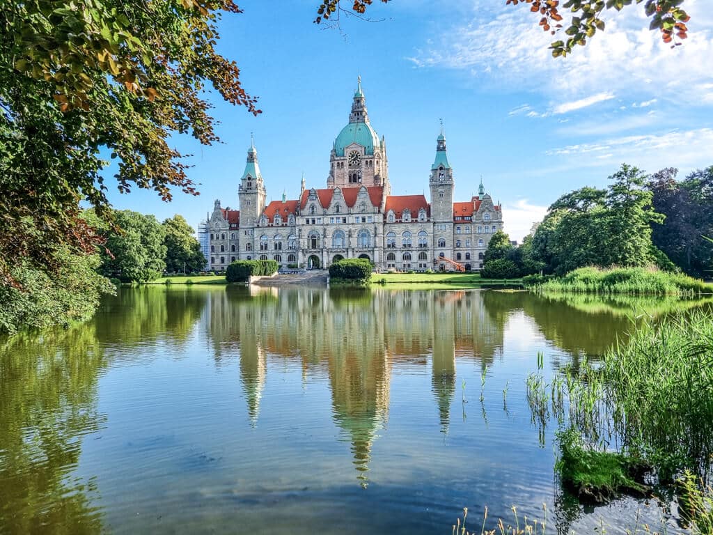 Ein großes, kunstvoll gestaltetes Gebäude mit einer grünen Kuppel und einem roten Dach spiegelt sich in einem ruhigen See. Das Bauwerk ist von üppigem Grün und Bäumen unter einem klaren blauen Himmel umgeben.
