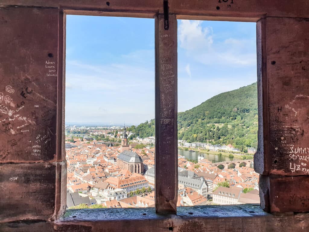 Blick durch ein Steinfenster auf eine Kirche und die Altstadt von Heidelberg mit dem Neckar