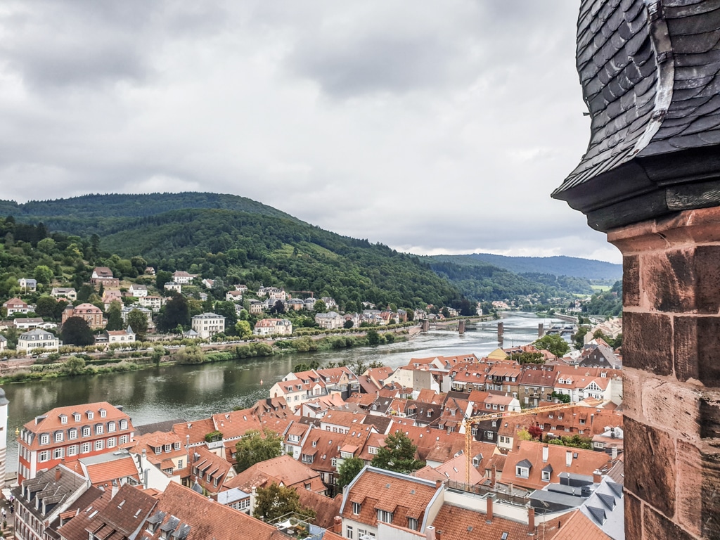 Blick über die Altstadt von Heidelberg, links davon der Neckar mit einer Brücke. Am linken Ufer einige Häuser und grün bewachsene Hügel dahinter